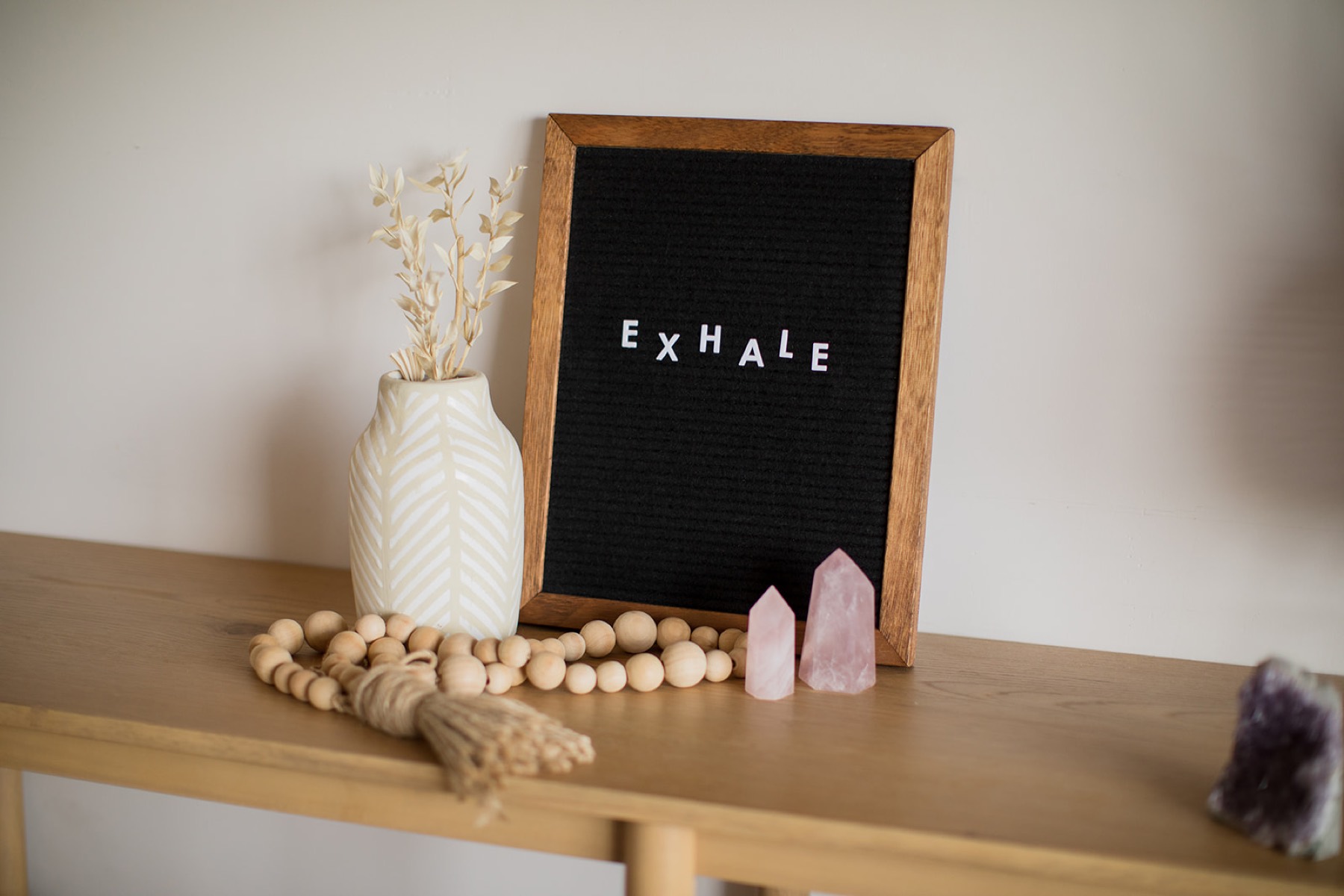 A wooden console styled with a letterboard reading 'Exhale', a vase of dried grasses, crystals, and wooden prayer beads